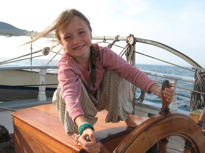 Nine-year-old Elinore Bailey, at the helm of a Tall Ship, foreshadows the fun to be had by families aboard SV Mystic in July (Photo Credit: Richard Bailey). 